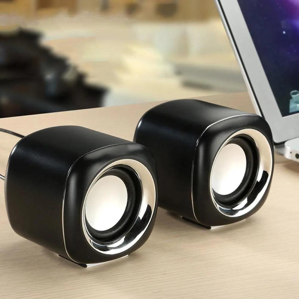 A pair of compact black or white wired desktop computer speakers with a volume scroll wheel.