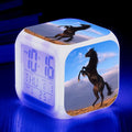 Small square LED alarm clock with colour-changing display on a bedside table.