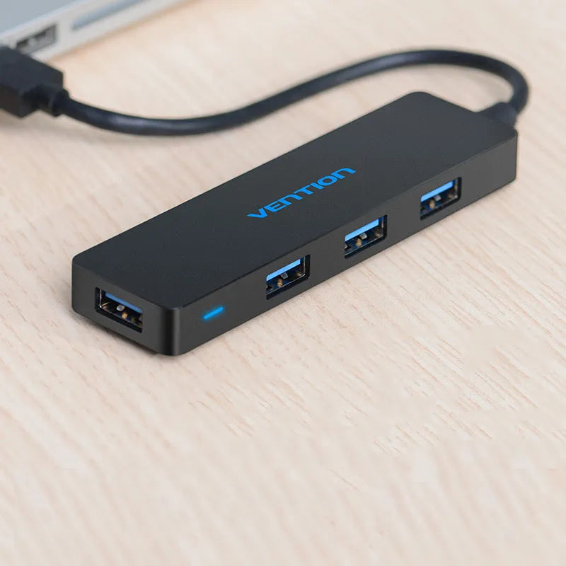 Black four-port USB 3.0 hub with a short cable on a desk next to a laptop