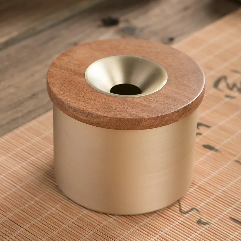 Brass-coloured copper ashtray with a wooden lid featuring a central hole, on a tabletop.