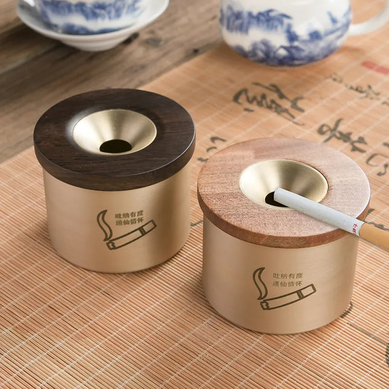Brass-coloured copper ashtray with a wooden lid featuring a central hole, on a tabletop.